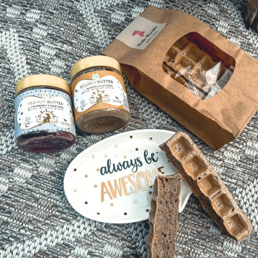 Baltic Biscuits Wuffel-Sticks in Papiertüte mit Sichtfenster, dazu Hundesnacks auf einem Teller mit der Aufschrift „always be awesome“ und zwei Gläser Hundeer Erdnussbutter in den Sorten Blaubeere & Hanfsamen sowie Banane & Leinsamen.