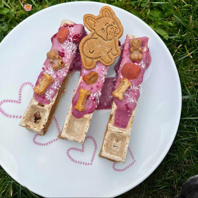 Drei Baltic Biscuits Wuffel-Sticks für Hunde auf einem Teller, dekoriert mit rosa Creme, Hundekeksen und einem Hundekeks in Form einer französischen Bulldogge.